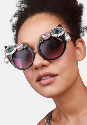 Lunettes de soleil en cat-eye avec montures noires, verres dégradés et décor embellissant comportant des perles et des pierres précieuses colorées. Accents dorés visibles.