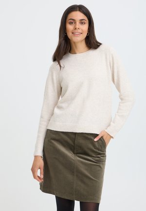 Jonge vrouw met lang donker haar draagt een crèmewitte trui, olijfgroene rok en zwarte panty's, glimlachend met één hand in haar zak.