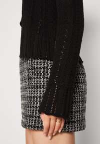 Femme portant un pull en maille noire avec une manche à motif et une jupe texturée noir et blanc, montrant une bague en or sur sa main gauche.