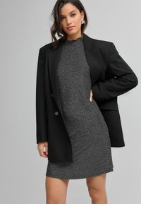 Čierny blazer na priliehavých, trblietavých šedých šatách so strapcovým výstrihom. Blazer má dvojité gombíky a uvoľnený siluetu.