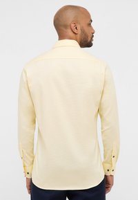 Camicia a maniche lunghe giallo chiaro con una sottile trama, polsini con bottone, e orlo dritto. Presenta un colletto standard e un dettaglio sul retro.