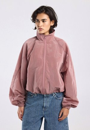 Jong persoon met kort donker haar, gekleed in een stofroze sweatvest met rits, ballonmouwen en hoge kraag, gecombineerd met een blauwe spijkerbroek.