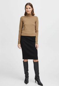 Maglione a dolcevita marrone chiaro, aderente, abbinato a una gonna a tubino nera a coste. La modella indossa stivali neri con tacco a ginocchio e punta affilata.