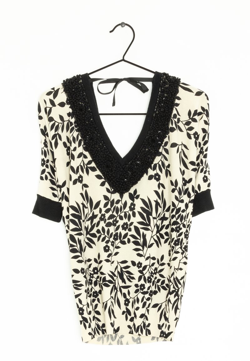 Blouse blanche à motif floral noir, col en V profond bordé d'une finition texturée noire, et manches courtes à revers noirs suspendue à un cintre noir.