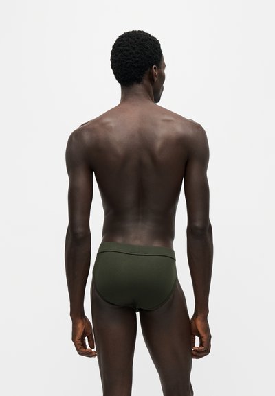 Modèle masculin à la peau foncée et aux cheveux bouclés, de dos, portant un slip vert foncé sur un fond clair uni.