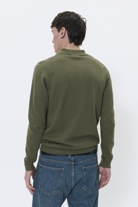 Camisa polo de manga larga verde oliva con cuello, que presenta una cintura ajustada y una tela texturizada. Jeans de mezclilla azul con bolsillos traseros visibles.