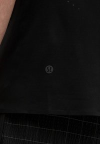 Čierne atletické tričko s hladkou textúrou, okrúhlym logom na dolnej prednej strane a jemnými perforáciami blízko výstrihu pre lepšiu priedušnosť.