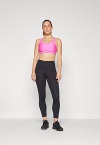Rozā sporta krūšturis ar baltu Under Armour logo, pārī ar augsto vidukli melnām legingām. Modelis valkā melnas sporta kurpes.
