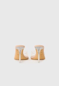 Une paire de sandales à talons hauts nudes avec des talons aiguilles transparents et des brides transparentes, photographiée de dos sur un fond blanc.