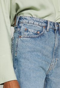 Vaqueros de mezclilla en azul claro con cintura alta, que cuentan con cinco bolsillos, costuras contrastantes y un acabado texturizado. Combinados con una blusa verde clara.