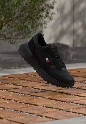 Tenis negru cu talpă groasă și logo roșu-alb-negru plutind deasupra plăcilor pătrate din lemn în aer liber.