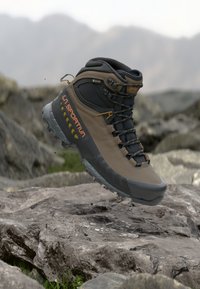 Bota de caminhada La Sportiva Gore-Tex em castanho e preto suspensa acima de terreno rochoso com um fundo de montanha desfocado.