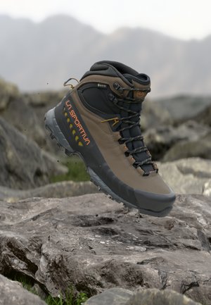 Bota de senderismo La Sportiva Gore-Tex en marrón y negro suspendida sobre un terreno rocoso con un fondo de montaña desenfocado.