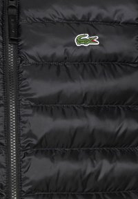 Svart puffer vest med horisontal quiltet tekstur, med en sentral glidelås og en liten grønn krokodillelogo på øverste venstre side.