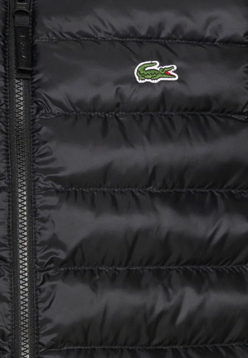 Gilet matelassé noir avec texture en quilting horizontal, présentant une fermeture éclair centrale et un petit logo de crocodile vert en haut à gauche.