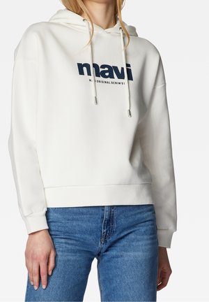 Personne portant un sweat à capuche blanc avec le texte « mavi » et un jean bleu clair, debout devant un fond uni.