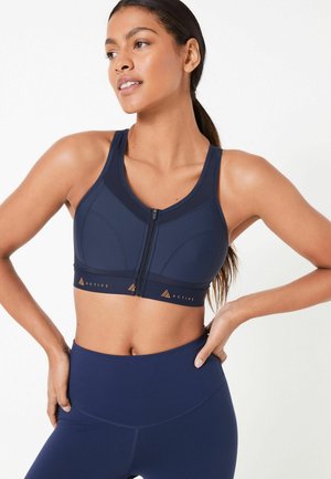 Navy sports bra with a front zipper, structured seams, and breathable mesh panels. Features an orange brand logo on the hem.