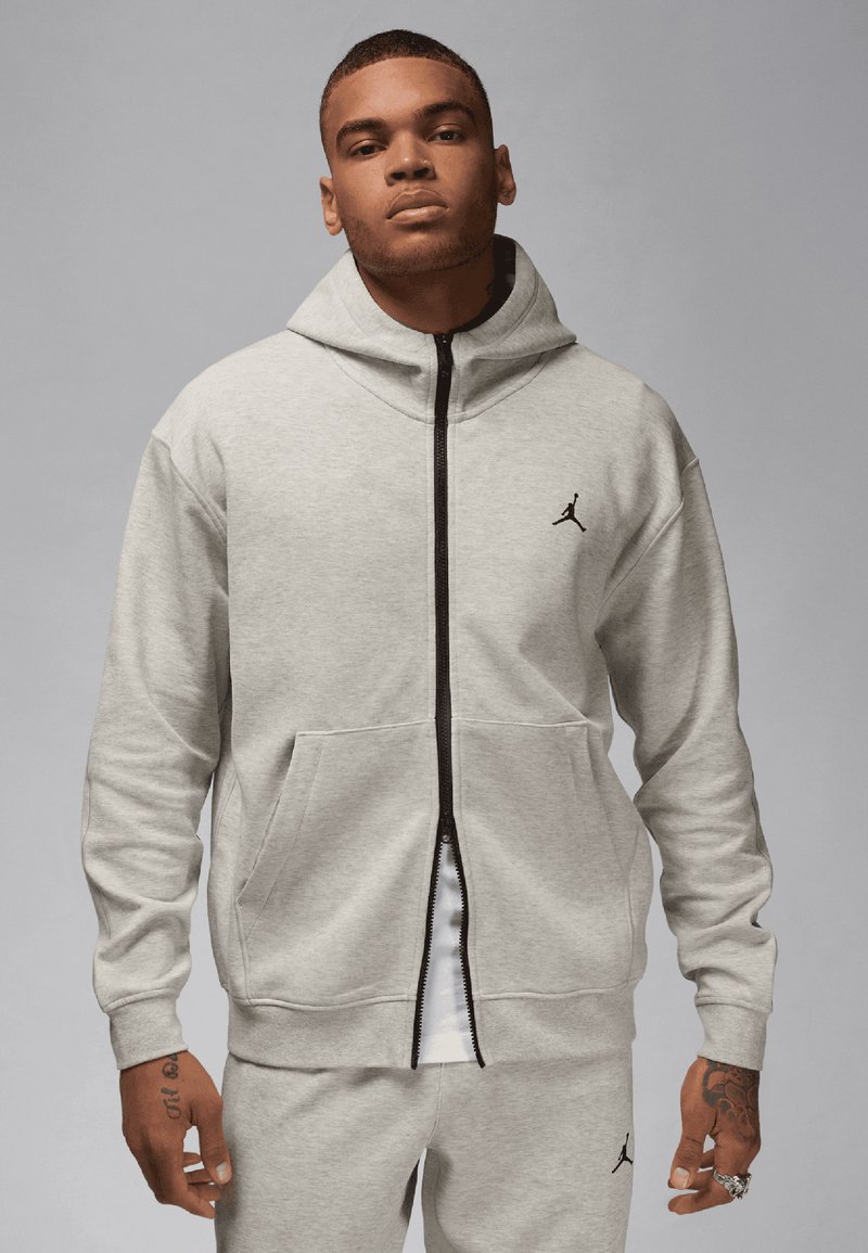 Szürke, cipzáros kapucnis pulóver puha anyagból, elülső kenguruzsebbel, magas gallérral és fekete Jumpman logóval a mellkasán.