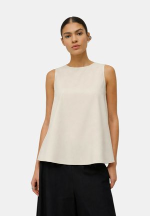 Donna con capelli scuri raccolti in uno chignon liscio, che indossa una canotta beige senza maniche e pantaloni neri larghi su uno sfondo chiaro e uniforme.