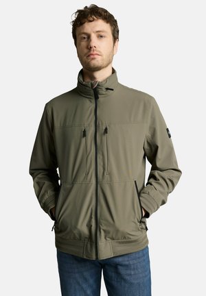 Mann trägt olivgrüne Jacke mit Reißverschluss und hohem Kragen sowie blaue Jeans, steht mit den Händen in den Taschen vor weißem Hintergrund.