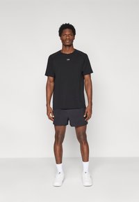 Μαύρο αθλητικό t-shirt με κοντά μανίκια και λογότυπο στο στήθος, συνδυασμένο με σκουρόχρωμο σορτς, λευκές κάλτσες στους αστραγάλους και λευκά αθλητικά παπούτσια.