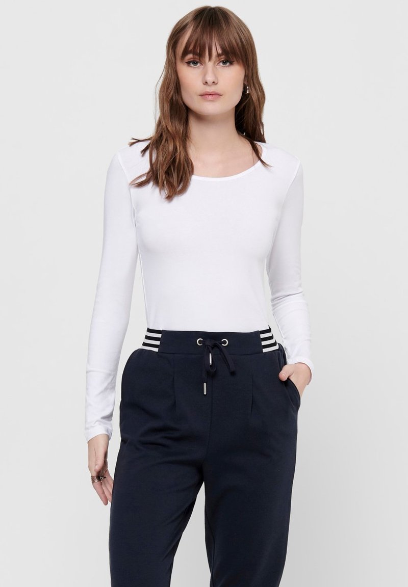 Langärmliges weißes Oberteil mit rundem Ausschnitt, kombiniert mit marineblauen Jogginghosen, die über einen Kordelzug und gestreifte Akzente am Bund verfügen.