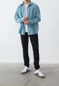 Camisa azul a rayas con un bolsillo en el pecho, camiseta blanca debajo, pantalones negros y zapatillas Adidas grises con detalles en negro y azul.