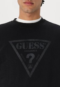 Sudadera negra con un gráfico triangular en gris oscuro que incluye el texto "GUESS® ORIGINALS", "WASHED" y un diseño de signo de interrogación.