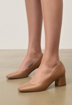 Nogi w beżowych skórzanych czółenkach na średnim obcasie, z zaokrąglonymi kwadratowymi noskami, stojące na gładkiej beżowej podłodze, na neutralnym tle.