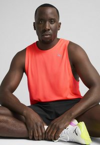 Sports tanktop i lys orange, med ærmeløst design og Puma-logo. Sorte shorts og hvide sportsko med neonlys.