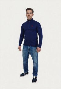 Marineblaue Zip-Knitpullover mit rotem Logo, getragen mit blauen Jeans und dunklen Sneakers. Mit gerippten Bündchen und Saum. Glatte Textur, taillierte Passform.