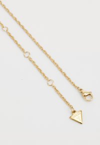 Gouden ketting met een gedraaid schakeldesign, een driehoekige hanger met de merknaam "GUESS," en een kreeftenhaaksluiting.