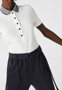 Polo shirt blanc à manches courtes avec un col rayé bleu marine, présentant une patte de boutonnage et un logo brodé. Associé à un pantalon taille haute bleu marine.