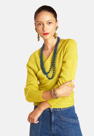 Yellow knitted sweater with long sleeves, paired with a blue beaded necklace and patterned bracelets, worn with denim jeans.