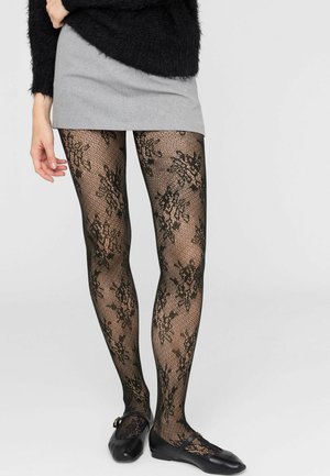 Beine mit schwarzen, blumigen Spitzenstrumpfhosen, einem grauen Minirock, einem schwarzen kuscheligen Pullover und schwarzen Mary Jane Ballerinas vor weißem Hintergrund.