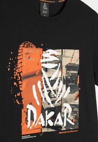 Czarna koszulka z nadrukiem w kolorze pomarańczowym i białym związanym z Rally Dakar, zawierająca pojazdy terenowe, współrzędne oraz stylizowany tekst i grafikę.