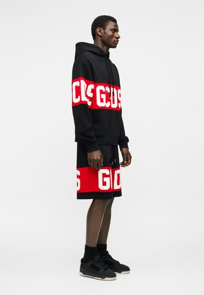 Uomo che indossa una felpa con cappuccio nera e pantaloncini con fasce orizzontali in rosso e bianco audaci, in piedi contro uno sfondo grigio chiaro uniforme.