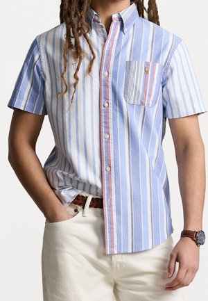 Man met lange dreadlocks die een blauw-wit gestreept, kort mouw, overhemdd met knopen draagt, met een gestreepte zak en witte broek.