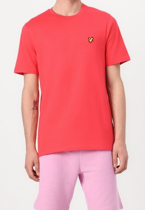 Mand iført en ensfarvet rød t-shirt med korte ærmer med et lille gult fuglelogo på brystet og lyserøde shorts.