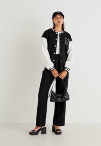 Veste varsity noir et blanc avec un patch de lettre, associée à un haut blanc, un pantalon noir et des sandales à plateforme noires et épaisses.
