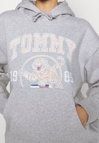 Sudadera gris de algodón suave con un gráfico en el frente. Presenta una ilustración de un tigre y el texto "TOMMY". Incluye una capucha con cordón y un bolsillo frontal.