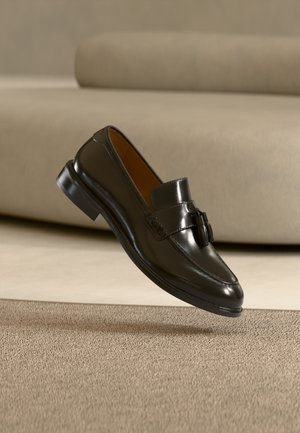 Black leather tassel loafer shoe floating above beige carpet with a beige sofa in the background.