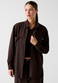 Braunes Hemd mit Knopfleiste und Tasche, weiche Textur und lockerer Schnitt, getragen über einem passenden dunklen Top, das ein minimalistisches Design betont.