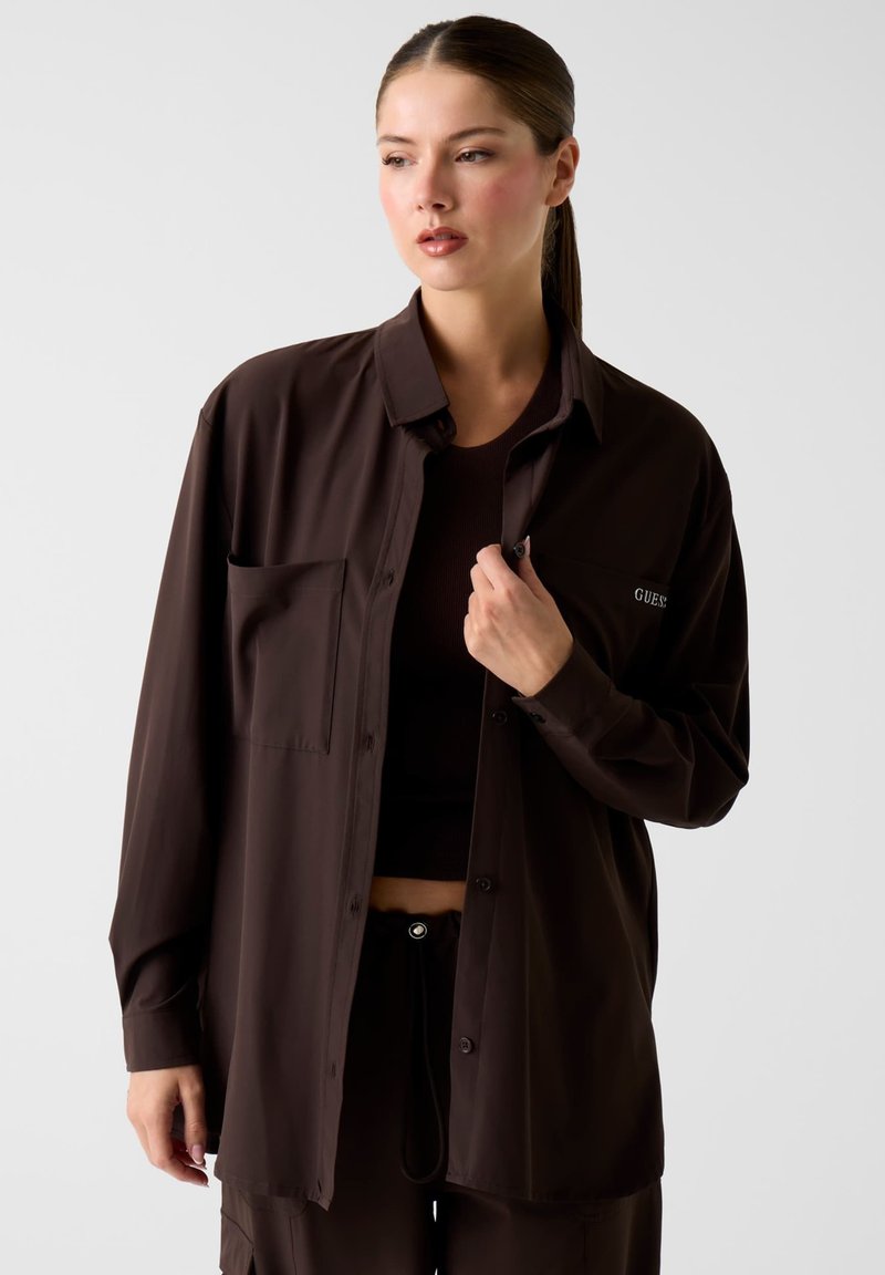 Braunes Hemd mit Knopfleiste und Tasche, weiche Textur und lockerer Schnitt, getragen über einem passenden dunklen Top, das ein minimalistisches Design betont.