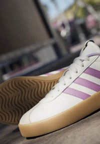 Zapatilla blanca con tres rayas moradas, parte superior de cuero texturizado y suela de goma. Presenta cordones redondos y un diseño de puntera redondeada.