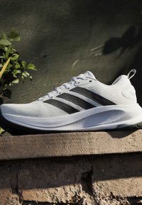 Chaussure de sport blanche avec des bandes noires, des lacets blancs et une semelle rembourrée, placée sur une surface en pierre texturée sur un fond sombre avec de la verdure.
