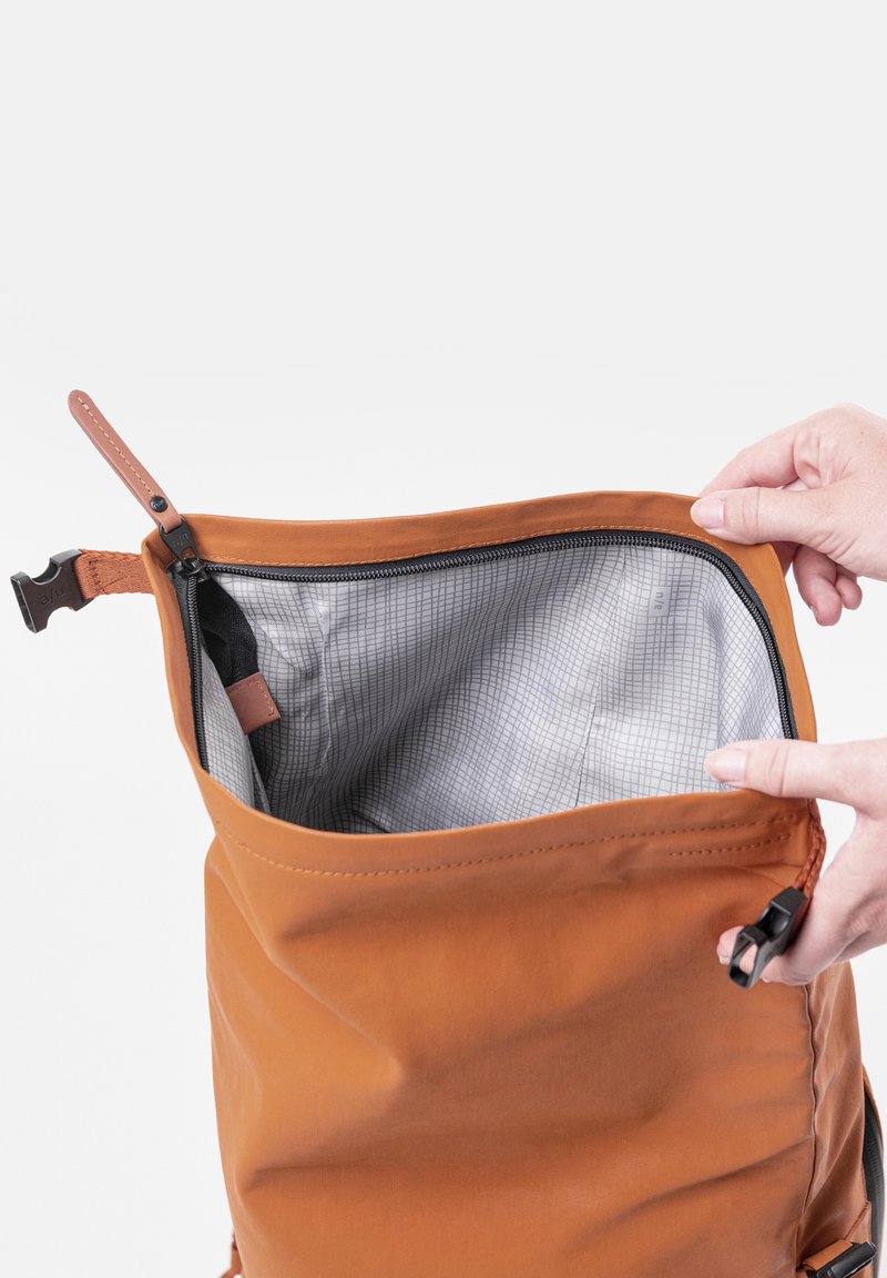 Borsa di stoffa arancione con apertura con zip, caratterizzata da un interno a motivo a griglia grigia. Una mano tiene la borsa, rivelando la sua texture e i dettagli del design.