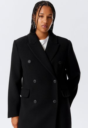 Woman with braided hair wearing a long black double-breasted coat over a white shirt, standing against a plain light background.