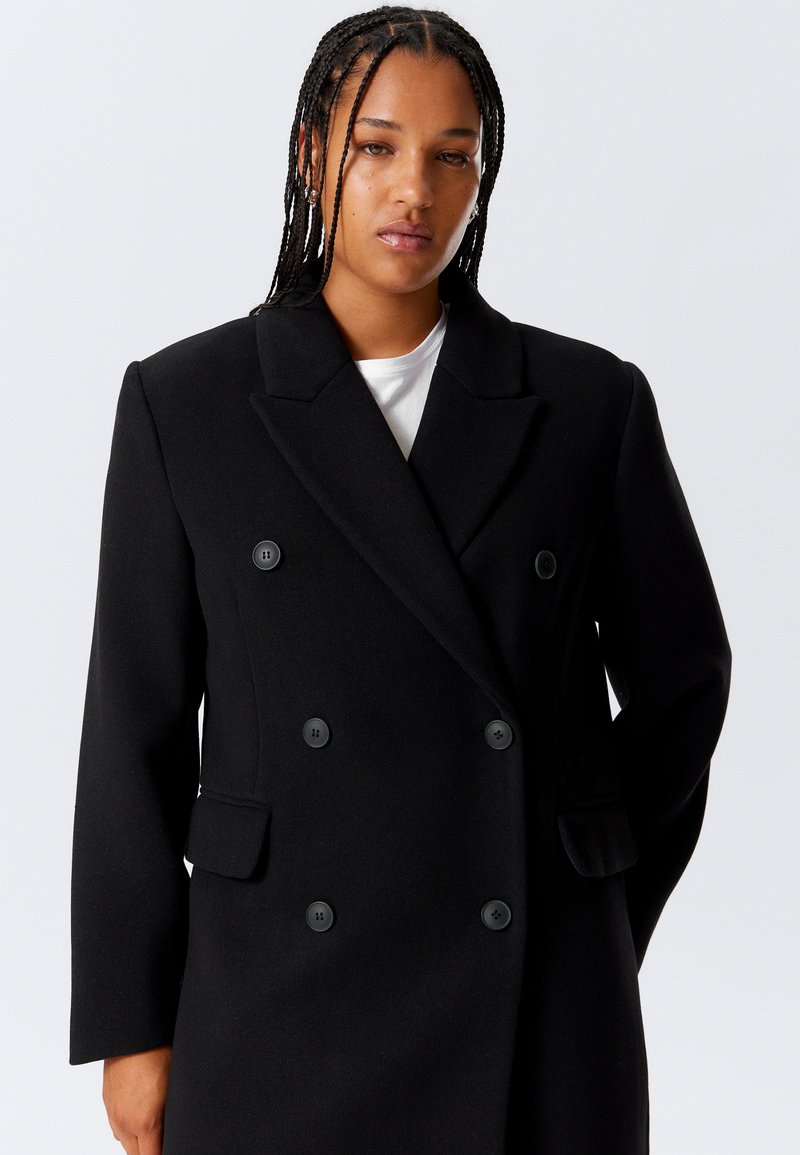 Femme avec des cheveux tressés portant un long manteau noir à double boutonnage sur une chemise blanche, se tenant devant un fond clair et uni.