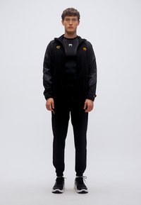 Μαύρο αθλητικό σετ φόρμας με φούτερ με φερμουάρ και κουκούλα, καθώς και εφαρμοστό μακρυμάνικο τοπ, με αντίθετες χρυσές λεπτομέρειες και υφασματόμορφο σχέδιο.